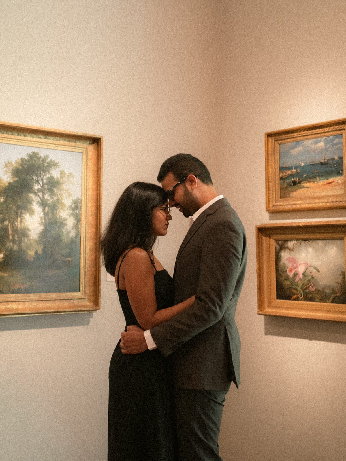 Couple embracing with their foreheads touching between framed landscape paintings, offering intimate engagement photo ideas for an indoor museum setting.