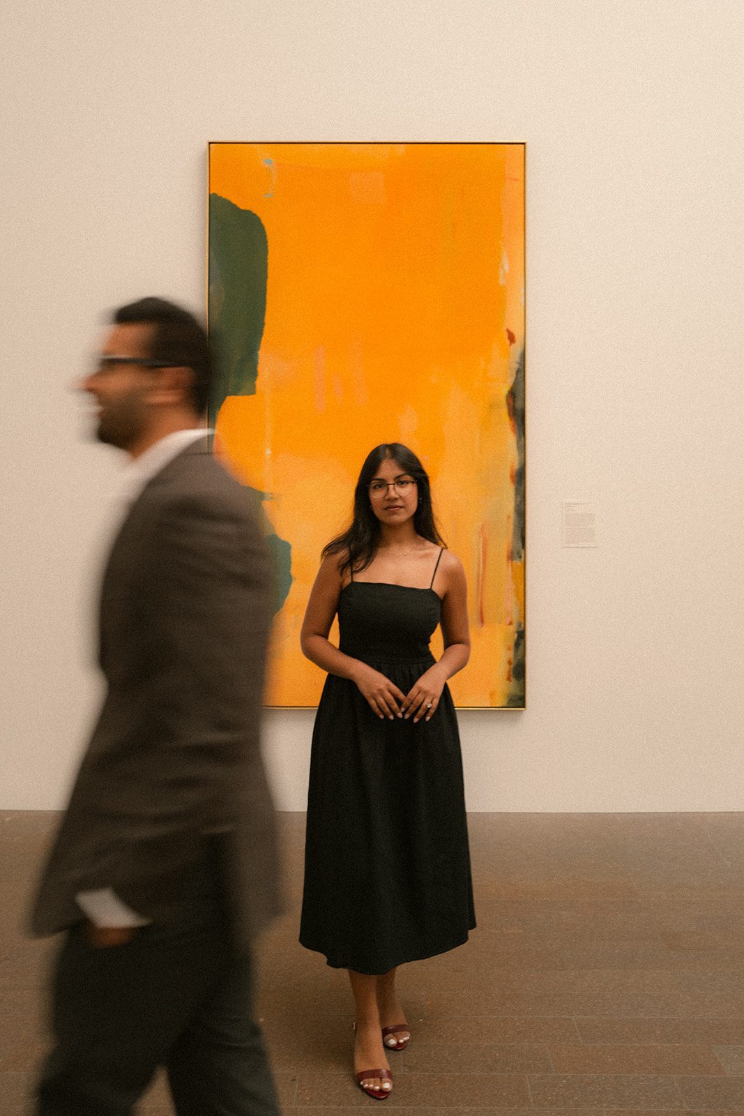 Woman in a black dress standing before a vivid orange abstract painting as her partner walks past, a creative composition for San Francisco Engagement Photos.