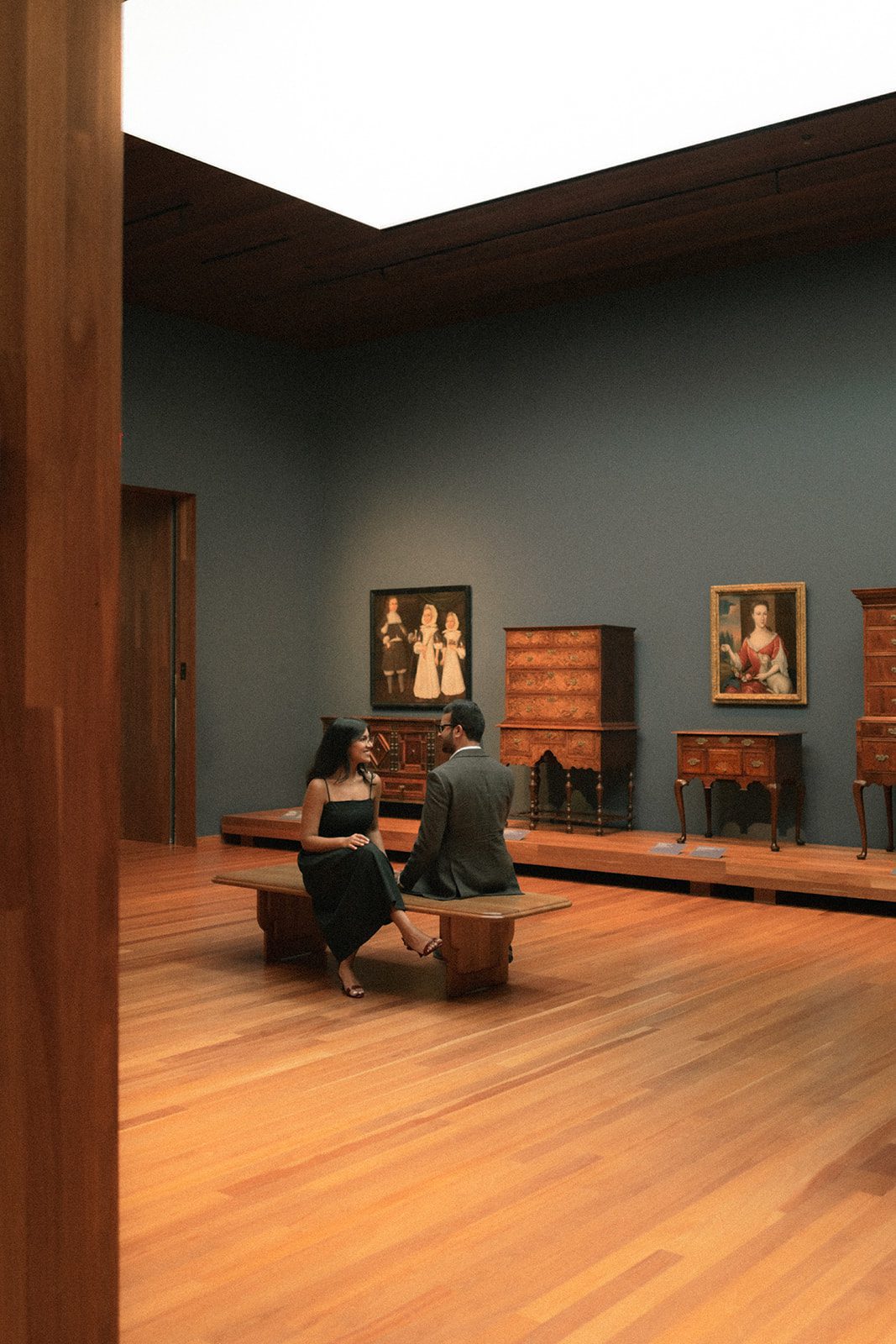 Couple sitting together on a wooden museum bench surrounded by antique furniture and portrait paintings, showcasing one of the more distinctive San Francisco engagement photo locations.