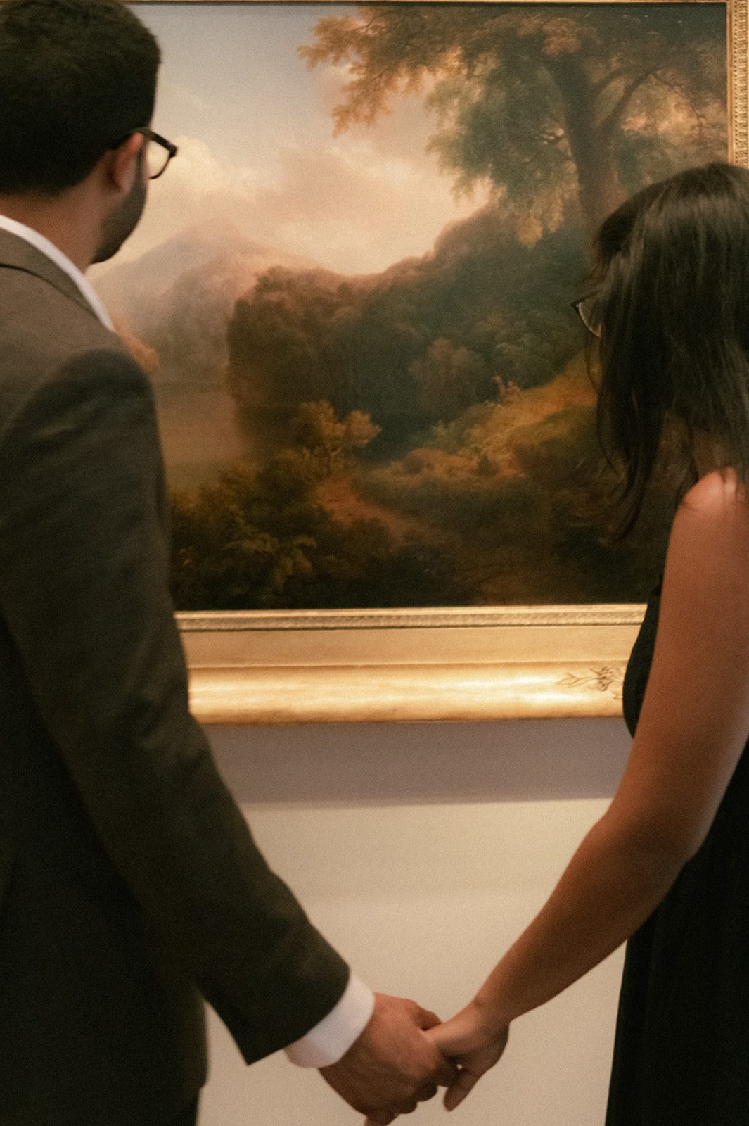 Couple holding hands while viewing a landscape painting during their San Francisco Engagement Photos, dressed in formal black and gray engagement photo outfits.