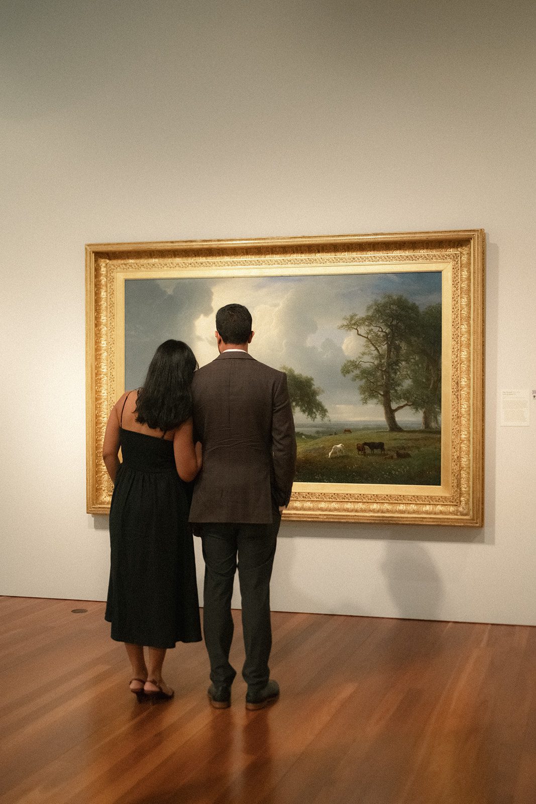 Couple standing shoulder to shoulder while viewing a framed landscape painting during their San Francisco engagement session in an art museum.