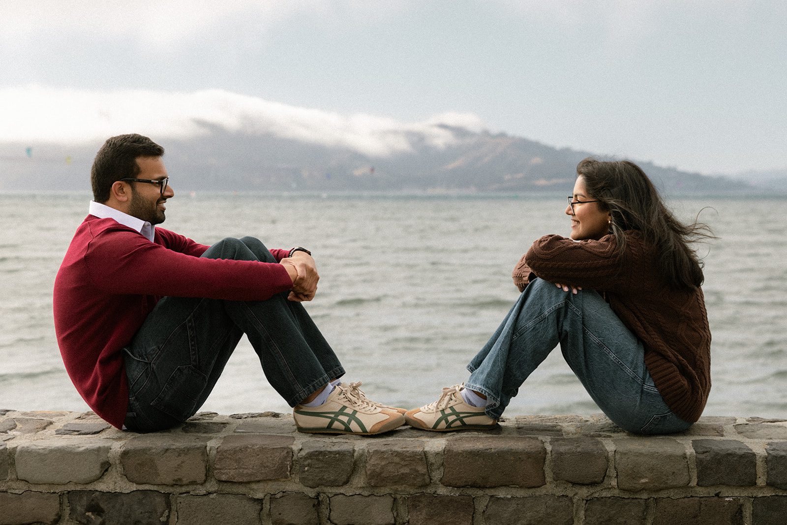 Couple sitting opposite each other on a stone waterfront wall with fog-covered hills across the bay, showcasing one of the scenic San Francisco engagement photo locations.