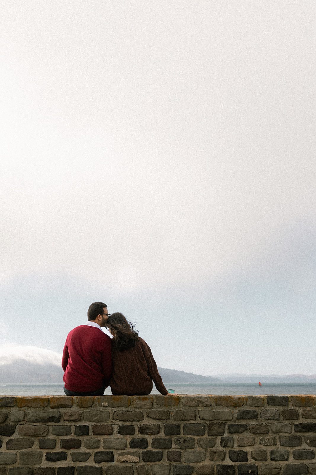 Couple sitting side by side on a stone wall overlooking the foggy bay, a quiet composition highlighting scenic San Francisco engagement photo locations.