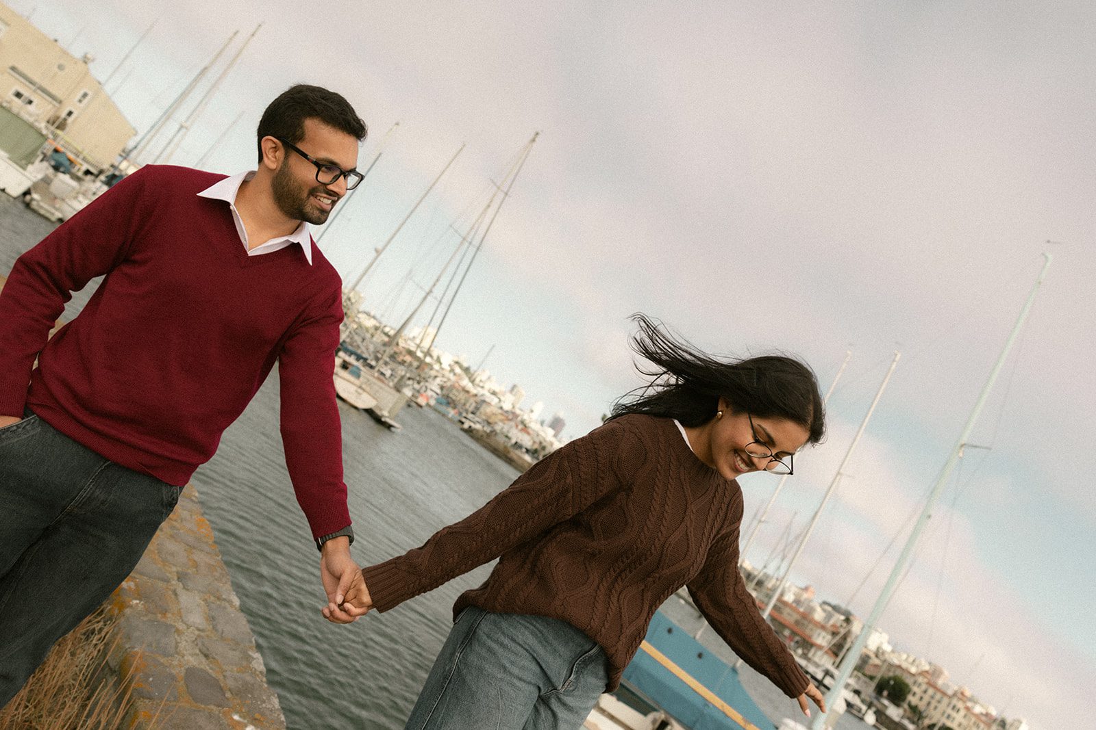 Couple holding hands while walking beside a marina in coordinated sweaters and jeans, showcasing relaxed engagement session outfits for engagement photos in San Francisco.