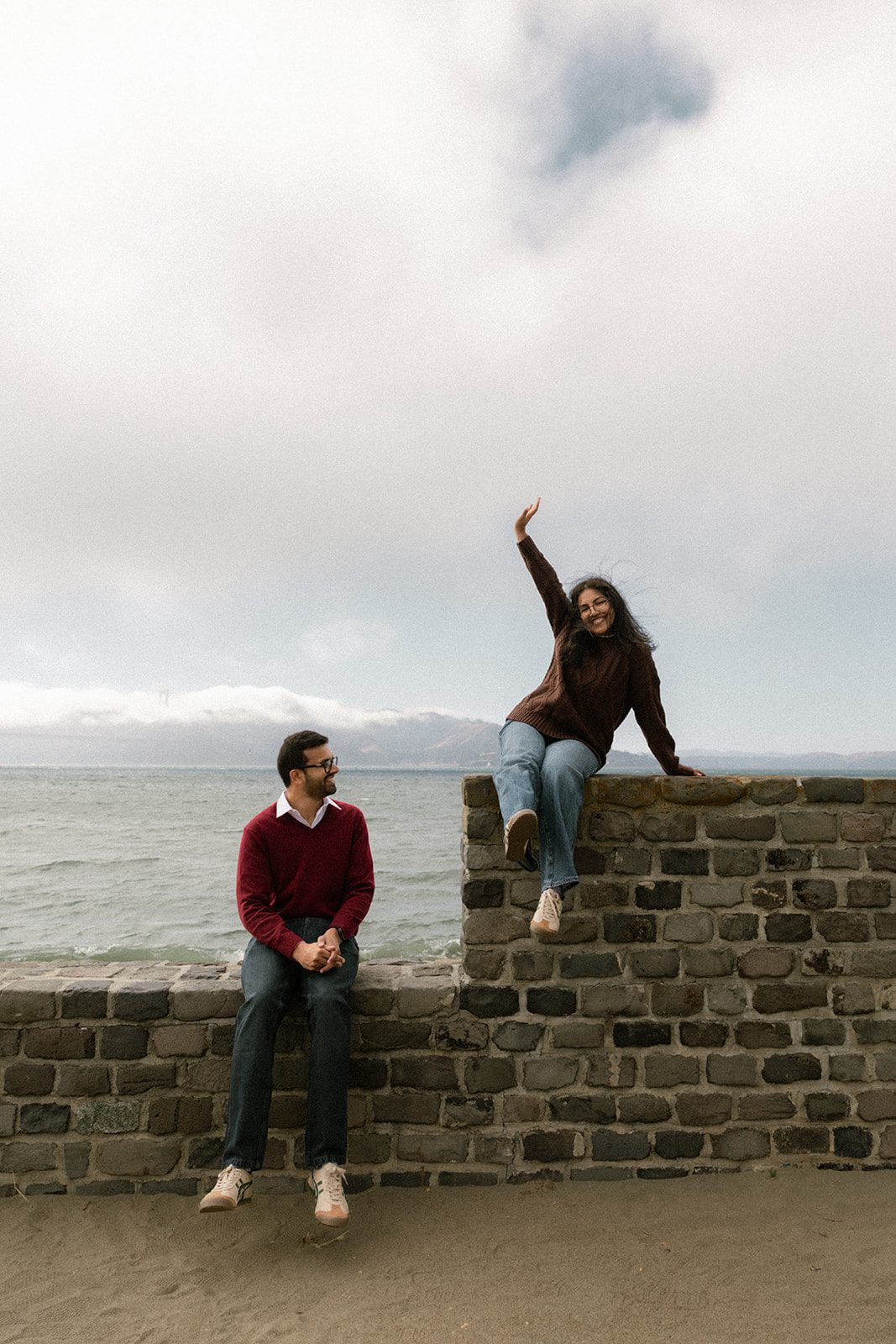 Couple playfully posing at different heights on a waterfront stone wall beneath a cloudy sky, bringing movement and personality to engagement photos in San Francisco.