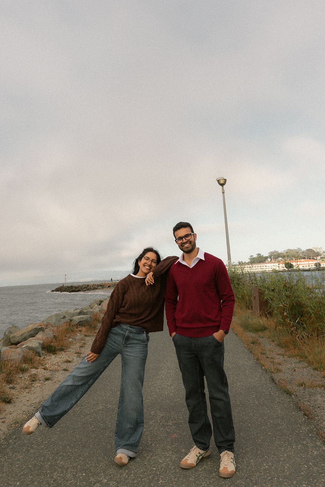 Couple posing together on a waterfront path in coordinated sweaters, jeans, and sneakers, offering relaxed engagement session outfits for San Francisco Engagement Photos.