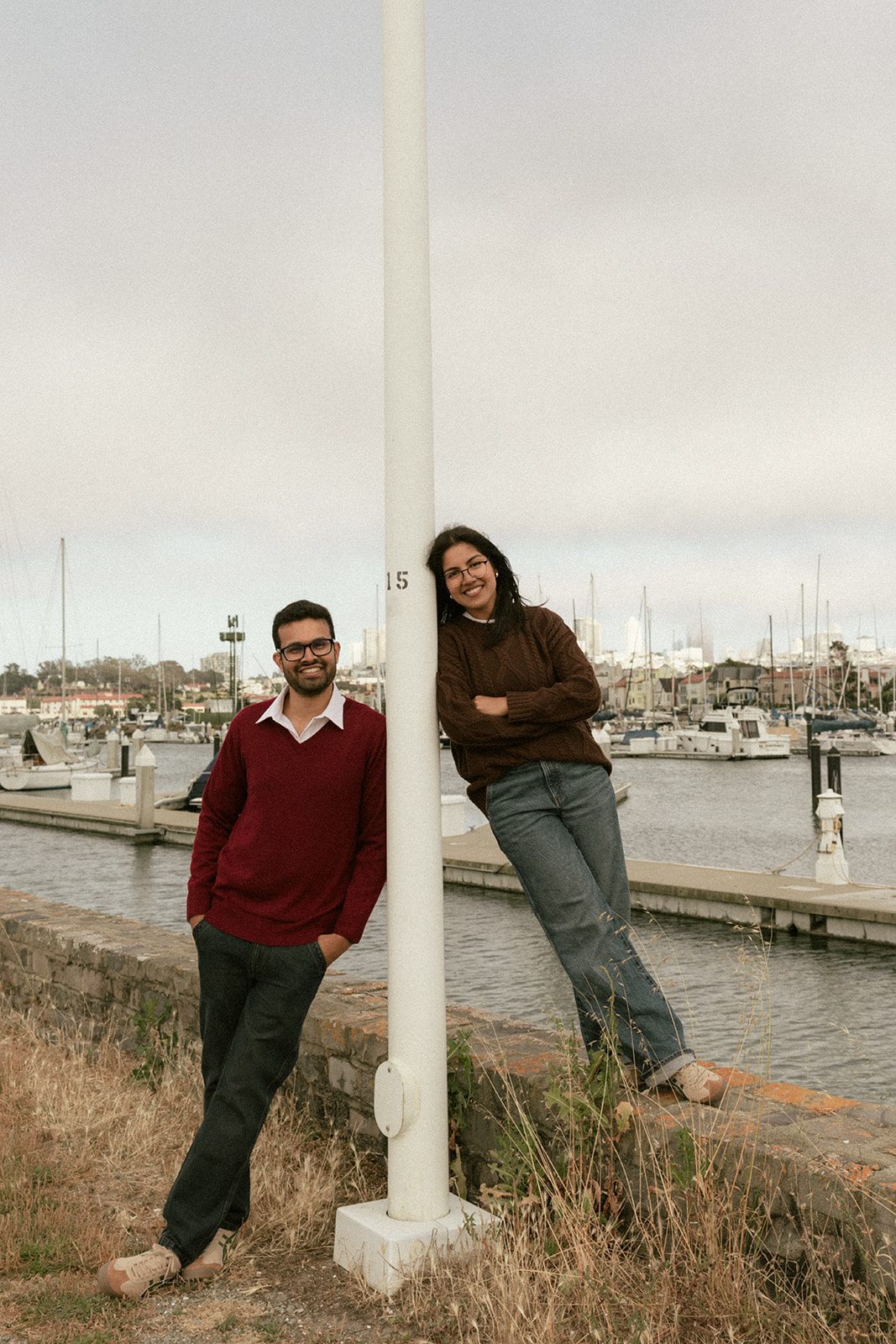 Couple posing on opposite sides of a marina light pole with sailboats behind them, a playful idea for San Francisco Engagement Photos in a waterfront setting.