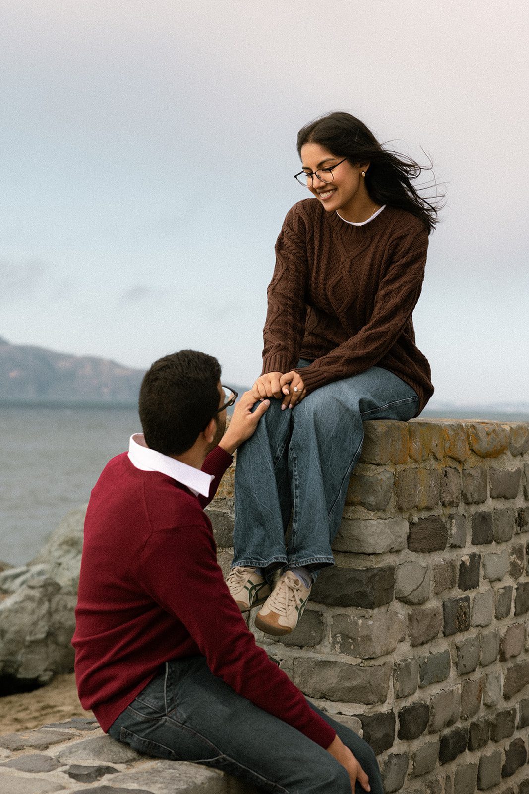 Couple holding hands at different levels of a stone seawall, showcasing relaxed engagement session outfits beside the San Francisco waterfront.