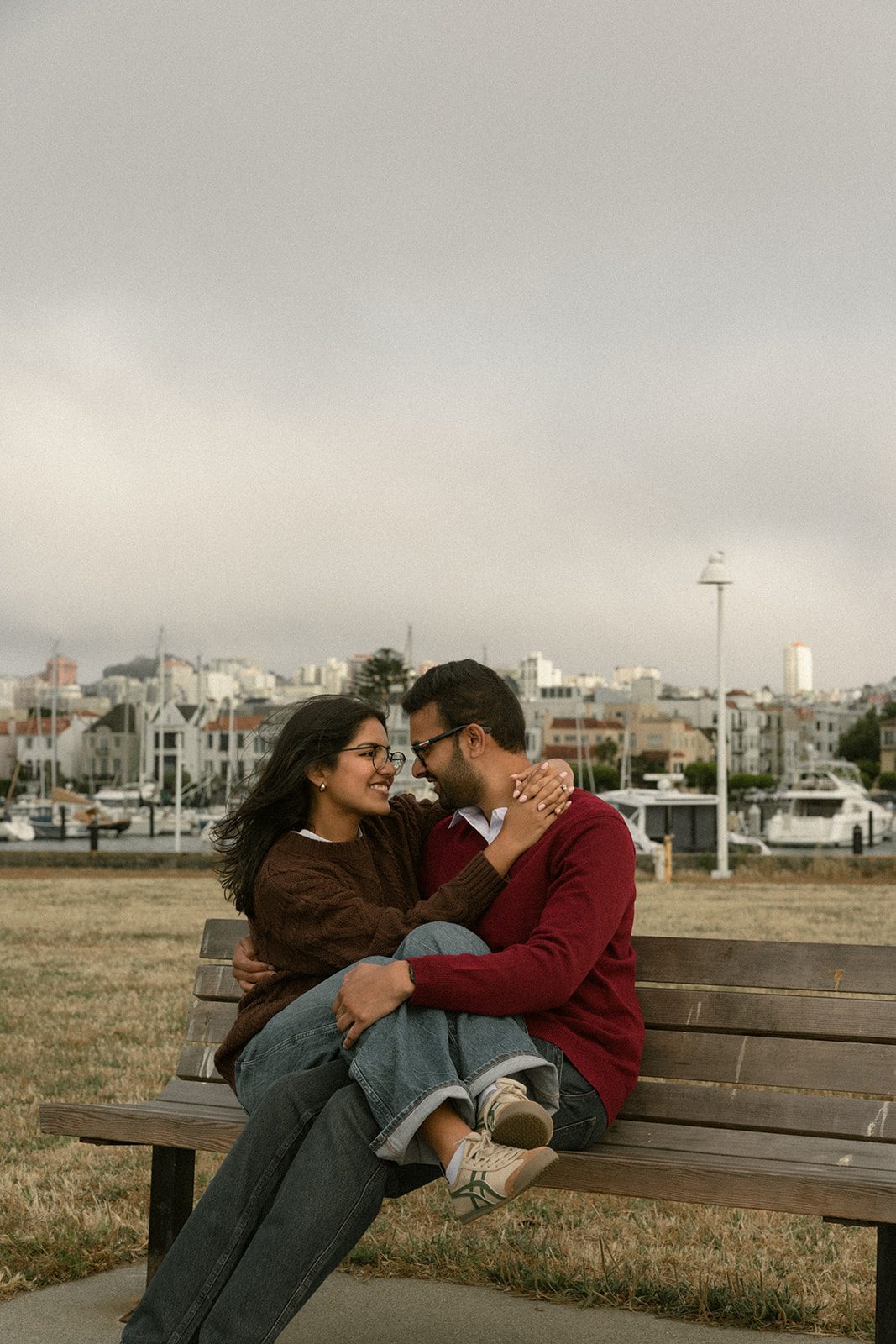 Couple cuddling on a park bench near a marina with San Francisco buildings in the background, a relaxed outdoor setting for engagement session ideas.