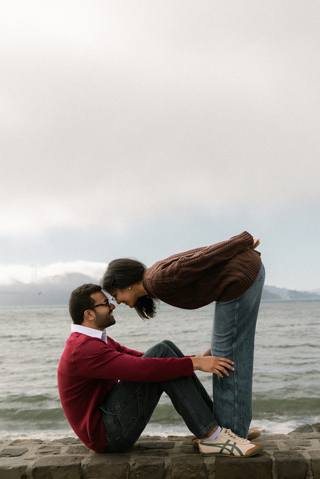 Couple leaning toward each other on a stone waterfront wall with foggy bay views, a playful pose for San Francisco Engagement Photos.