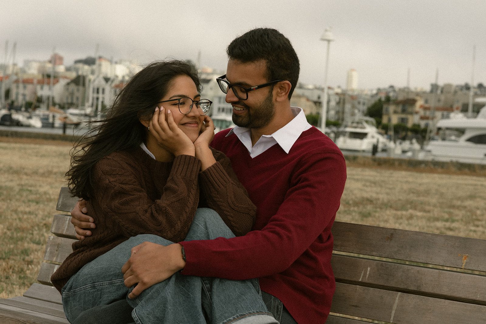 Couple cuddling on a waterfront bench with boats and city buildings behind them, a casual engagement photo idea for a San Francisco engagement session.