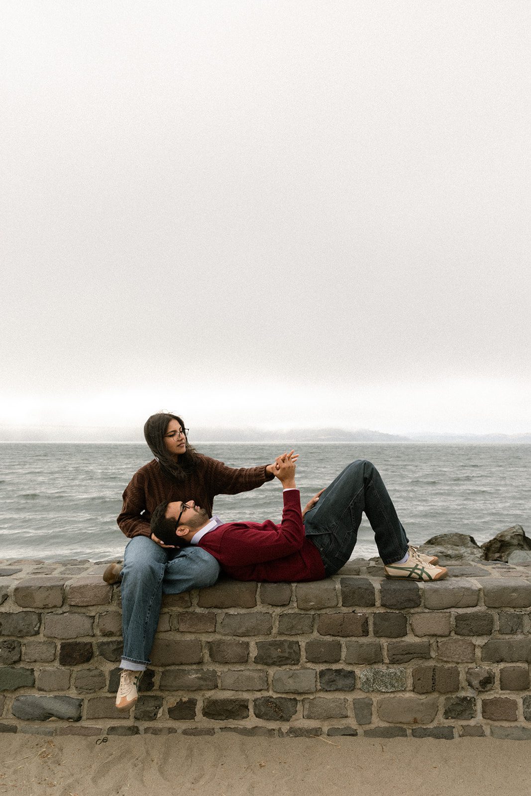 Couple relaxing on a stone seawall with one partner resting across the other’s lap, a casual engagement photo idea with foggy bay views.