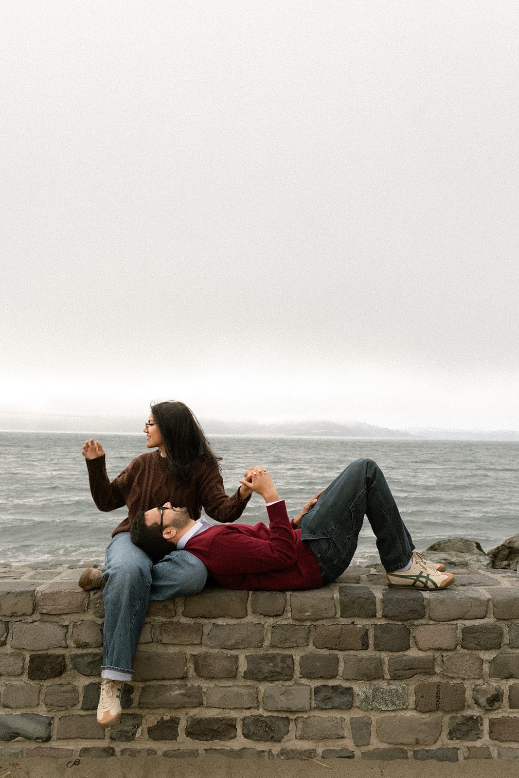 Couple relaxing on a stone waterfront wall during a casual San Francisco engagement session, wearing sweaters and jeans with foggy bay views behind them.