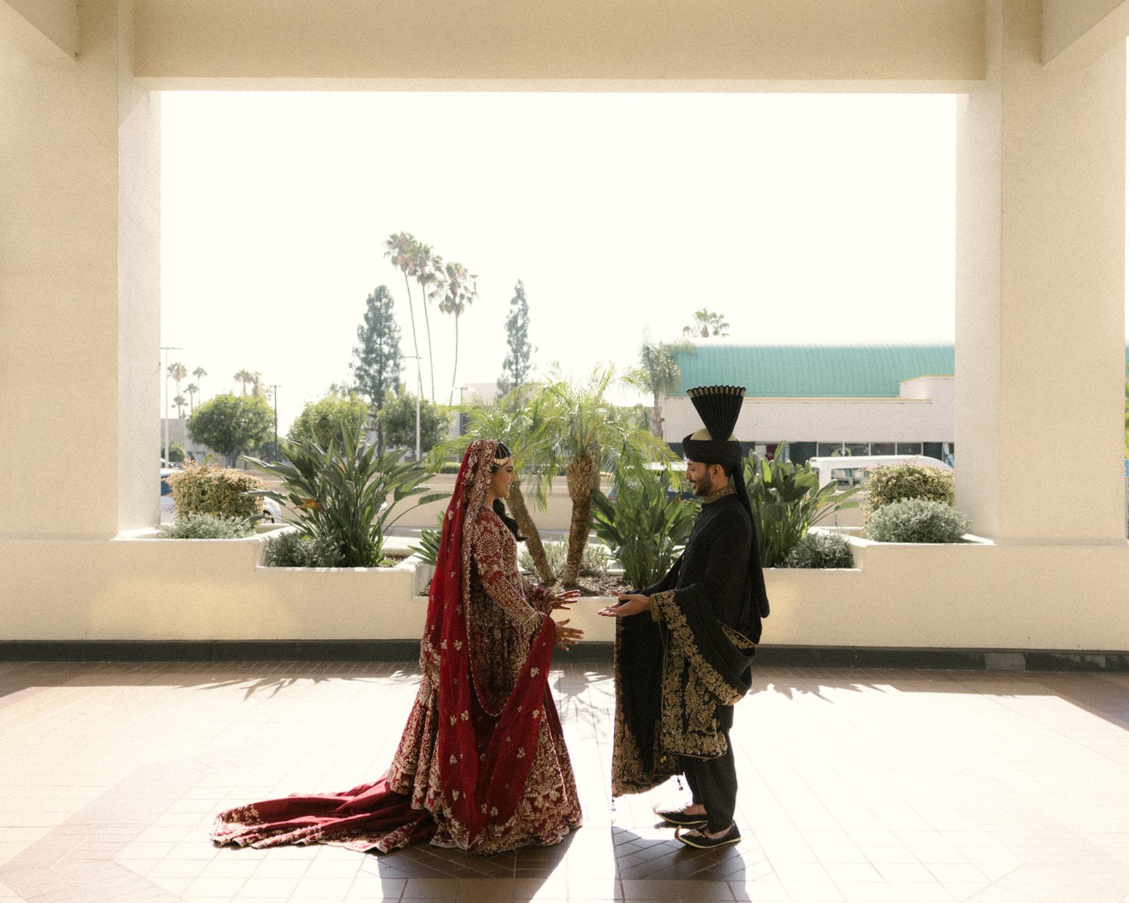 Bride and groom facing one another in red and black traditional attire during their indian wedding, framed by bright architecture and palm trees.