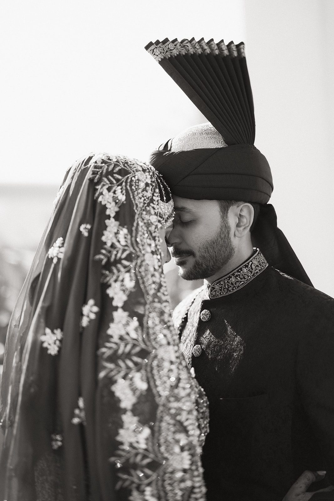 Black-and-white close-up of a bride and groom touching foreheads in ornate traditional attire during their indian wedding, highlighting the embroidered veil and groom’s turban.