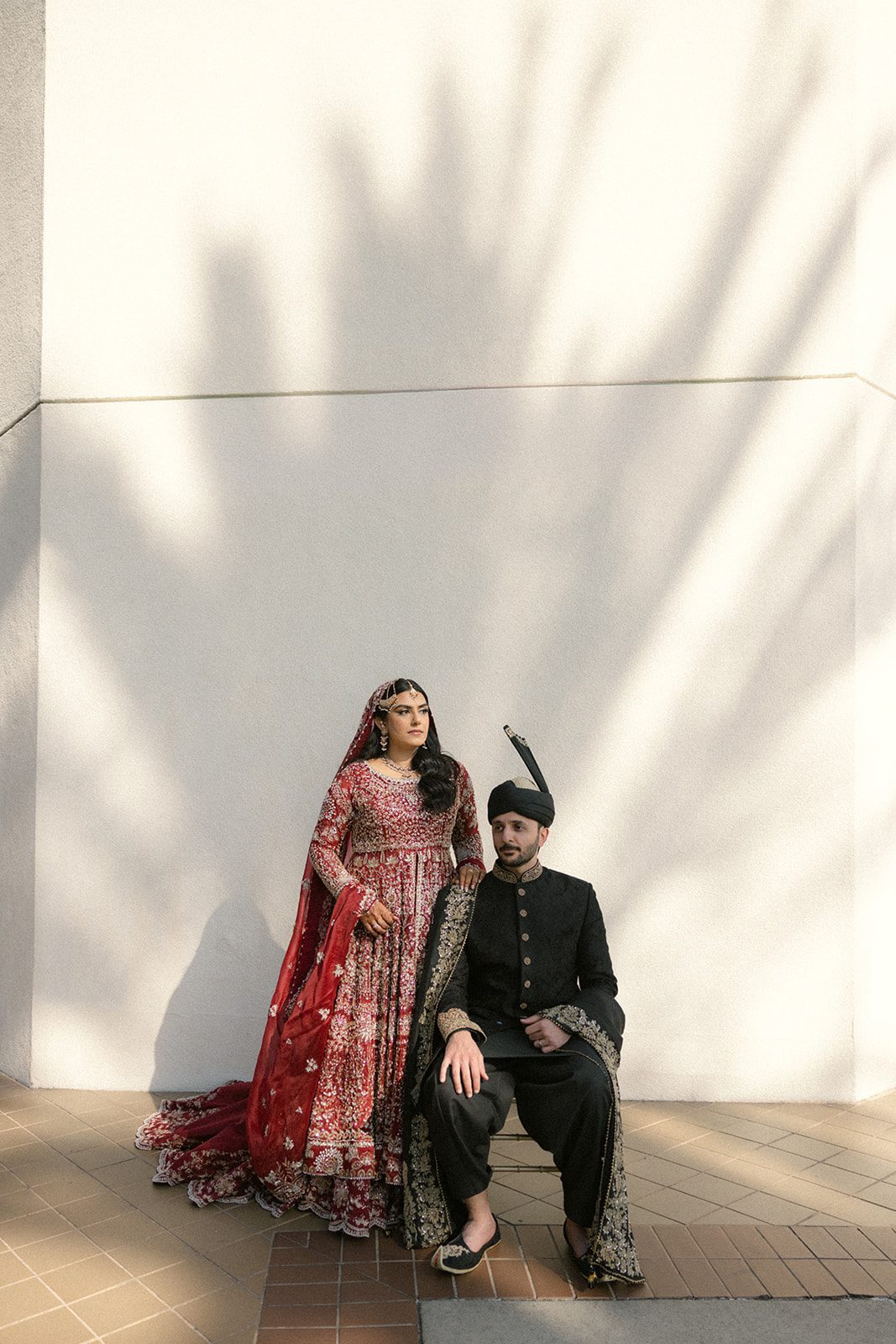 Bride in a red embroidered lehenga standing beside the seated groom in black traditional attire for a modern south asian wedding portrait against a sunlit wall.