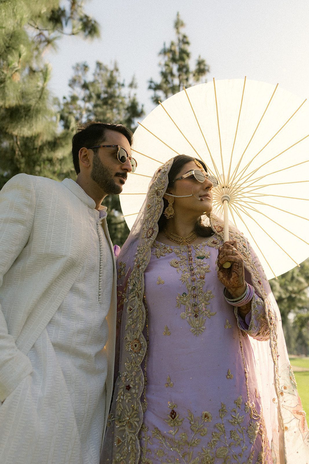 Couple in traditional attire and sunglasses posing beneath a white parasol during their outdoor south asian wedding, photographed in warm natural light by an indian wedding photographer.