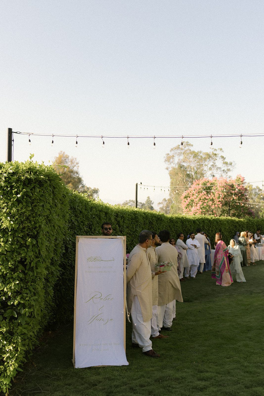 Guests gathering on a garden lawn beside the welcome sign before an outdoor indian wedding ceremony under string lights.