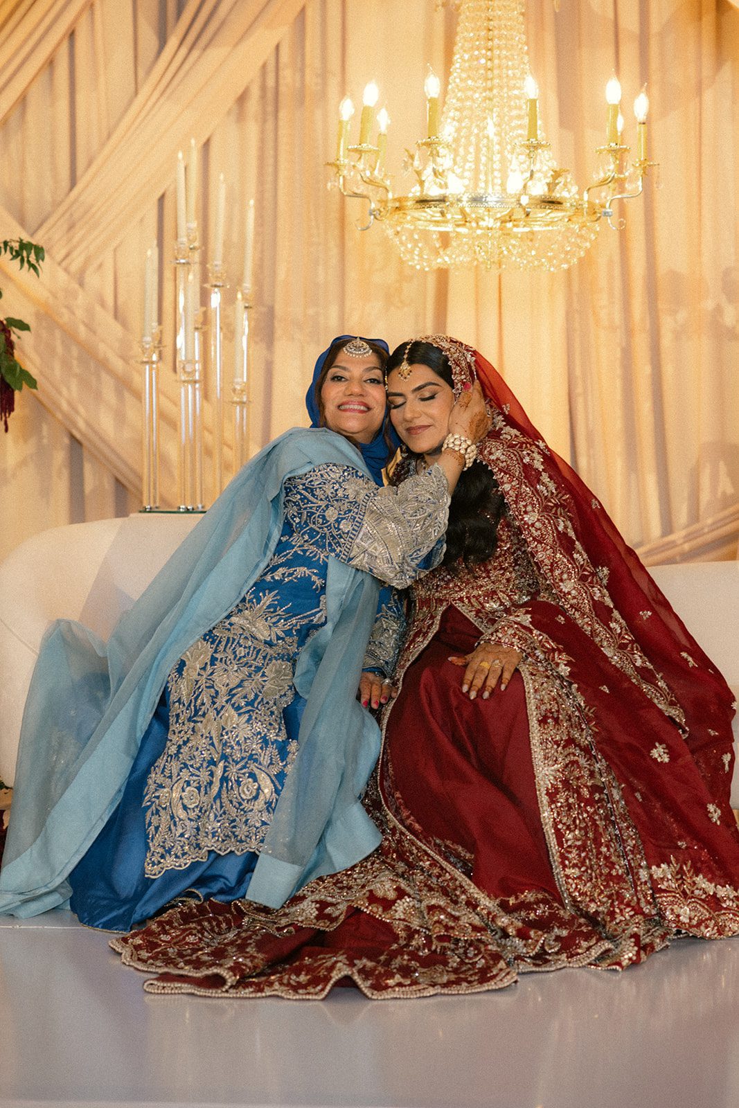Bride in a red embroidered outfit seated with a woman in blue traditional attire during a south asian wedding celebration.