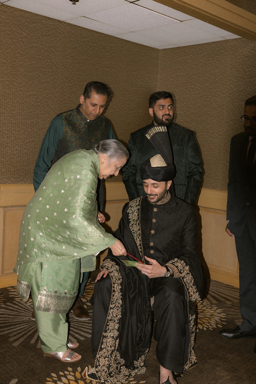 Groom in a black embroidered sherwani and turban receiving help from family before a multi day wedding ceremony.