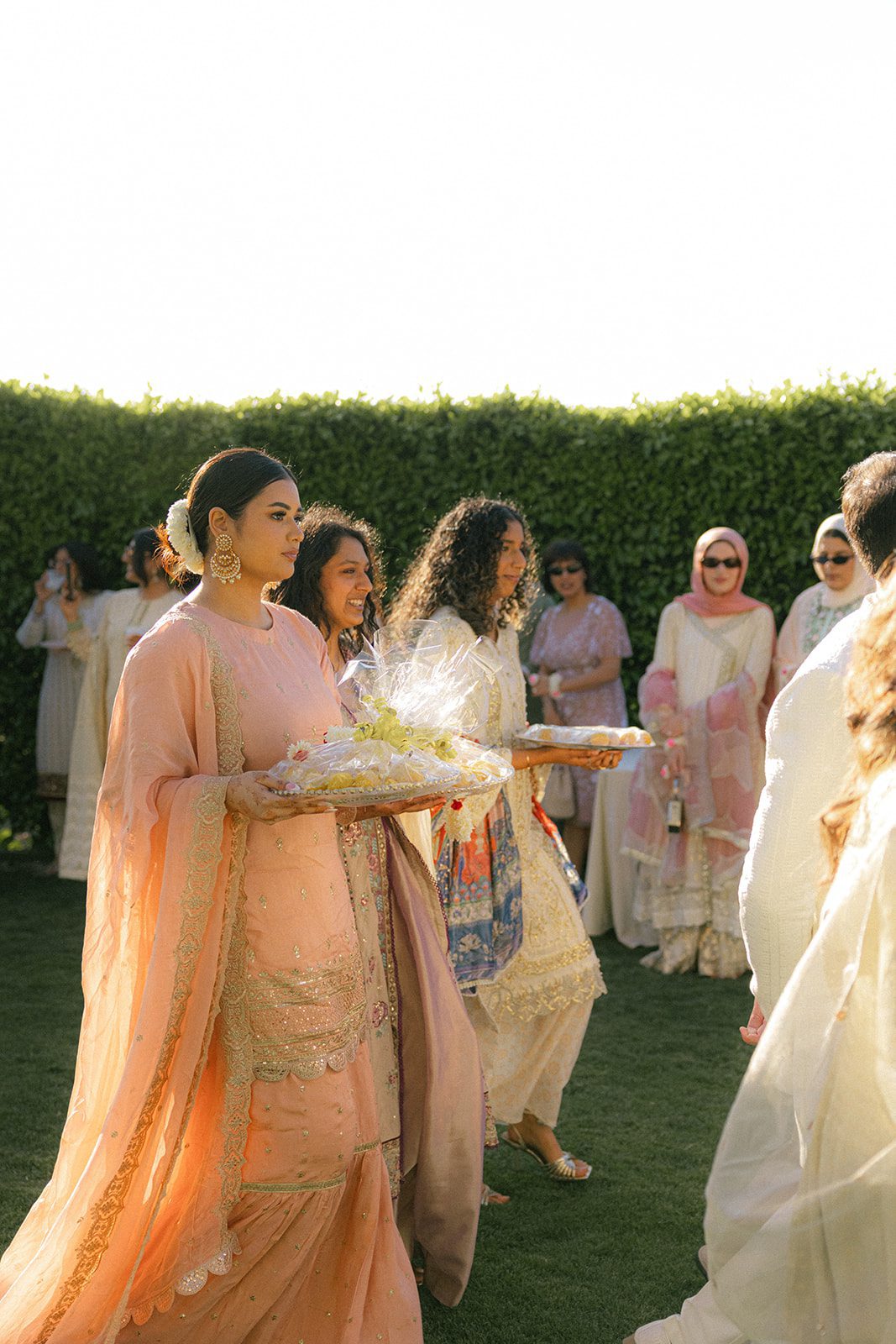 Women in pastel traditional attire carrying ceremonial trays during a sunlit south asian wedding procession on the lawn.