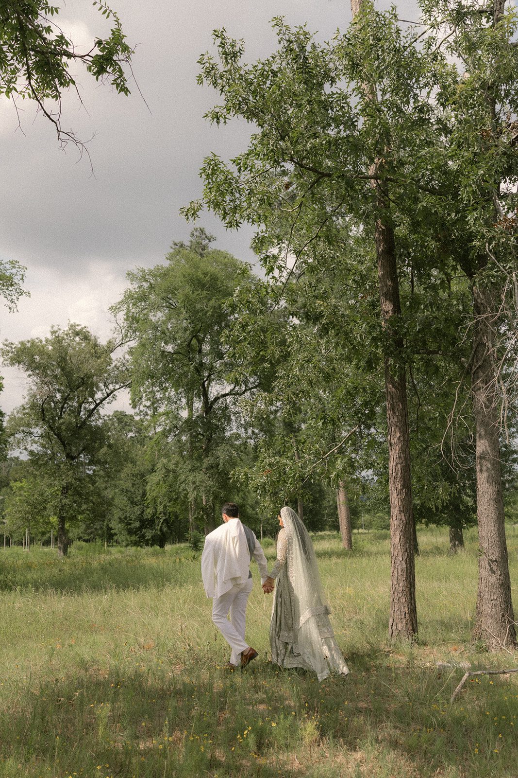 Newlyweds holding hands as they walk through a grassy woodland in traditional attire, a quiet portrait from their multi day wedding celebration.