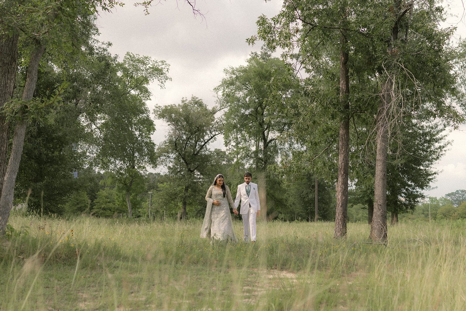 Bride and groom holding hands while walking through a grassy wooded landscape after their indian wedding ceremony.
