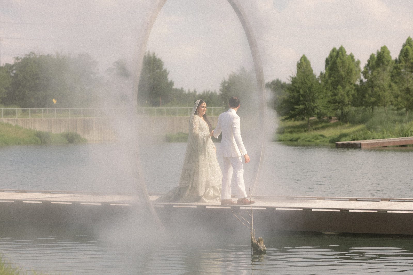 Bride and groom walking hand in hand beside a lake and misting sculpture, captured by an indian wedding photographer during their outdoor portraits.