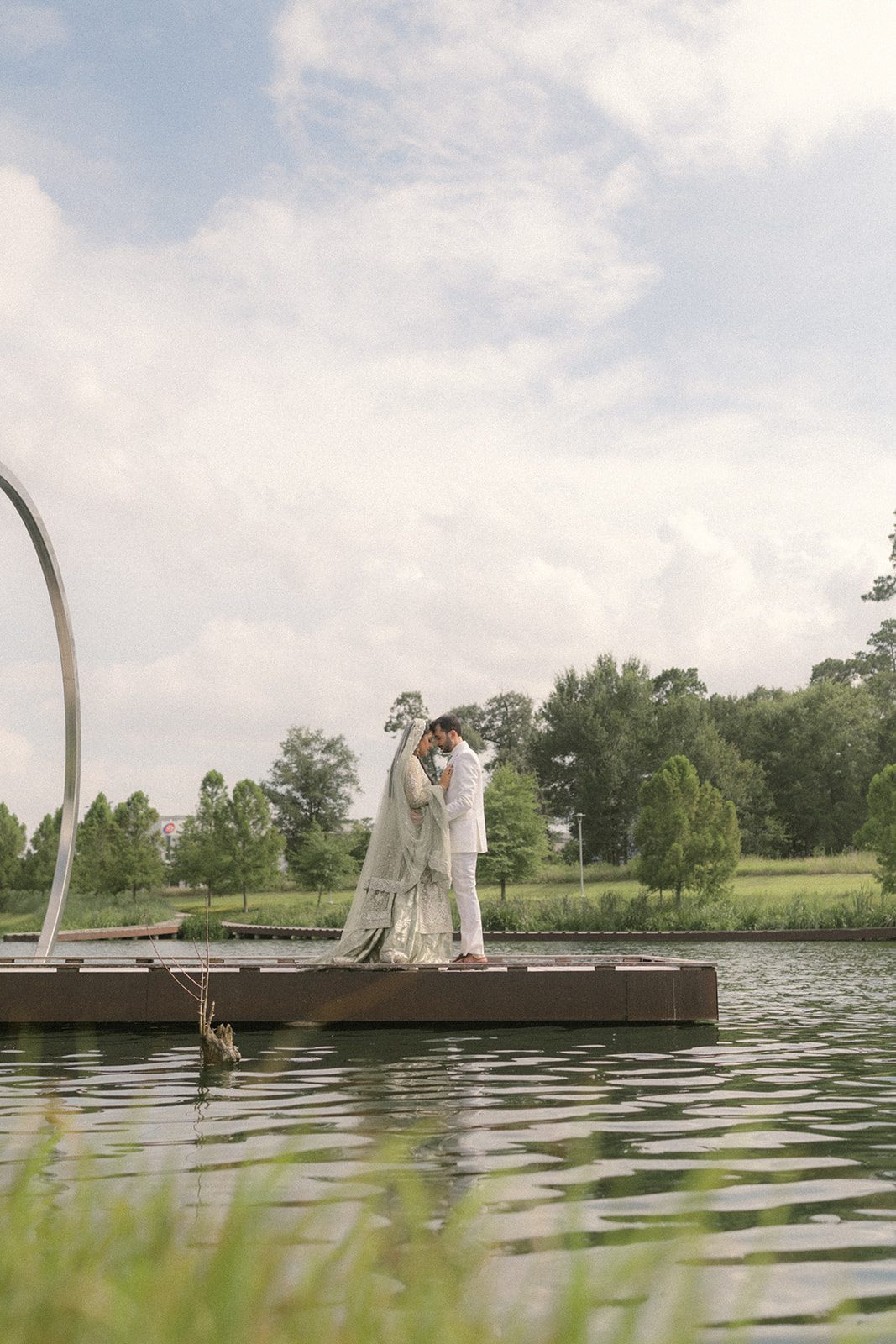Newlyweds in traditional wedding attire standing together on a dock beside a landscaped pond, a quiet portrait from their multi day wedding celebration.