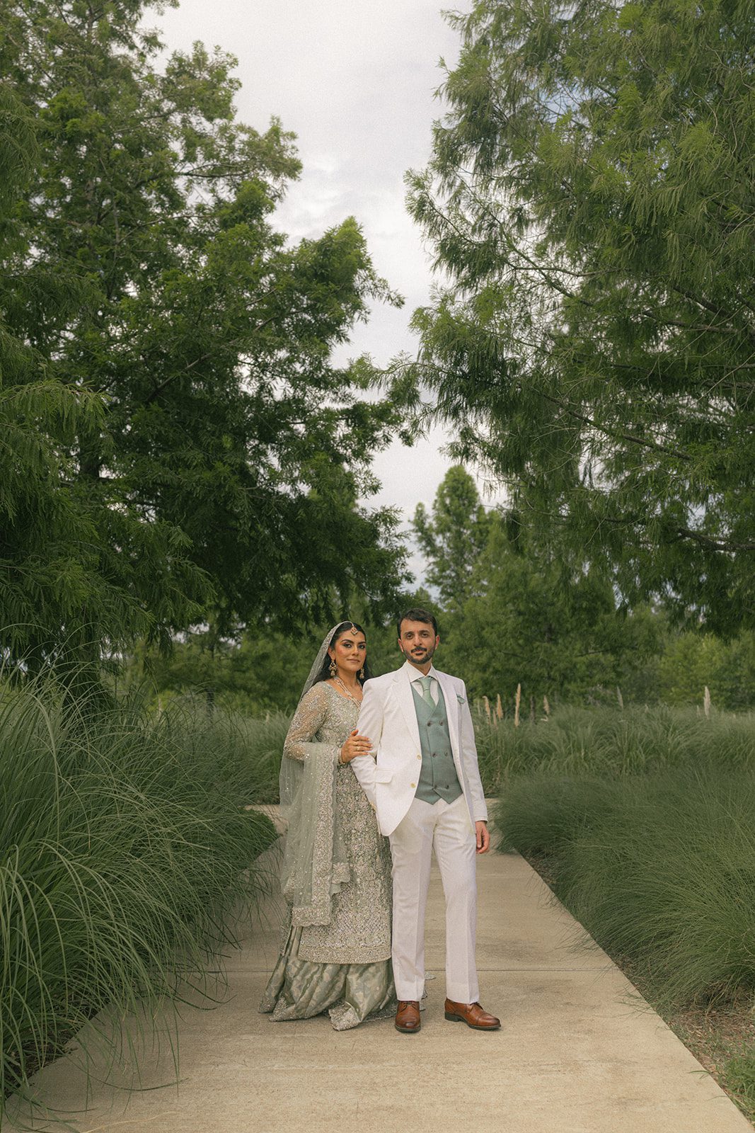 Couple posing along a tree-lined garden path in silver and white attire, photographed outdoors during their south asian wedding.