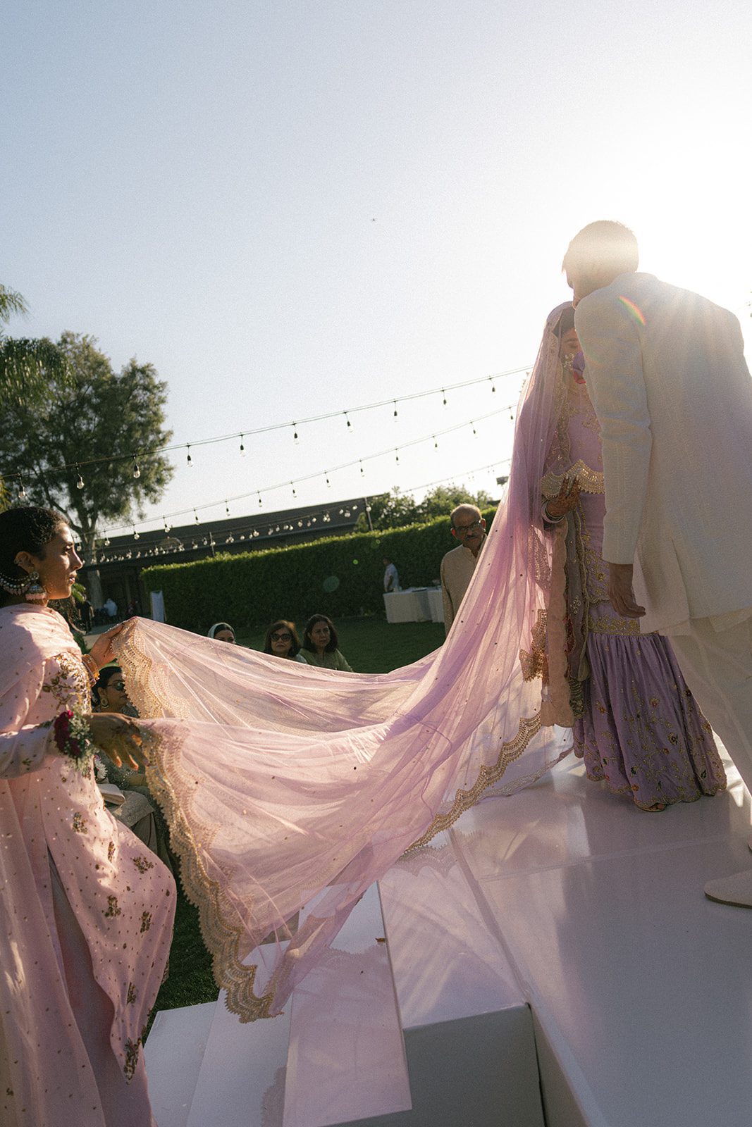 Bride in lavender embroidered attire approaching the groom as a family member holds her flowing veil during an outdoor multi day wedding ceremony.