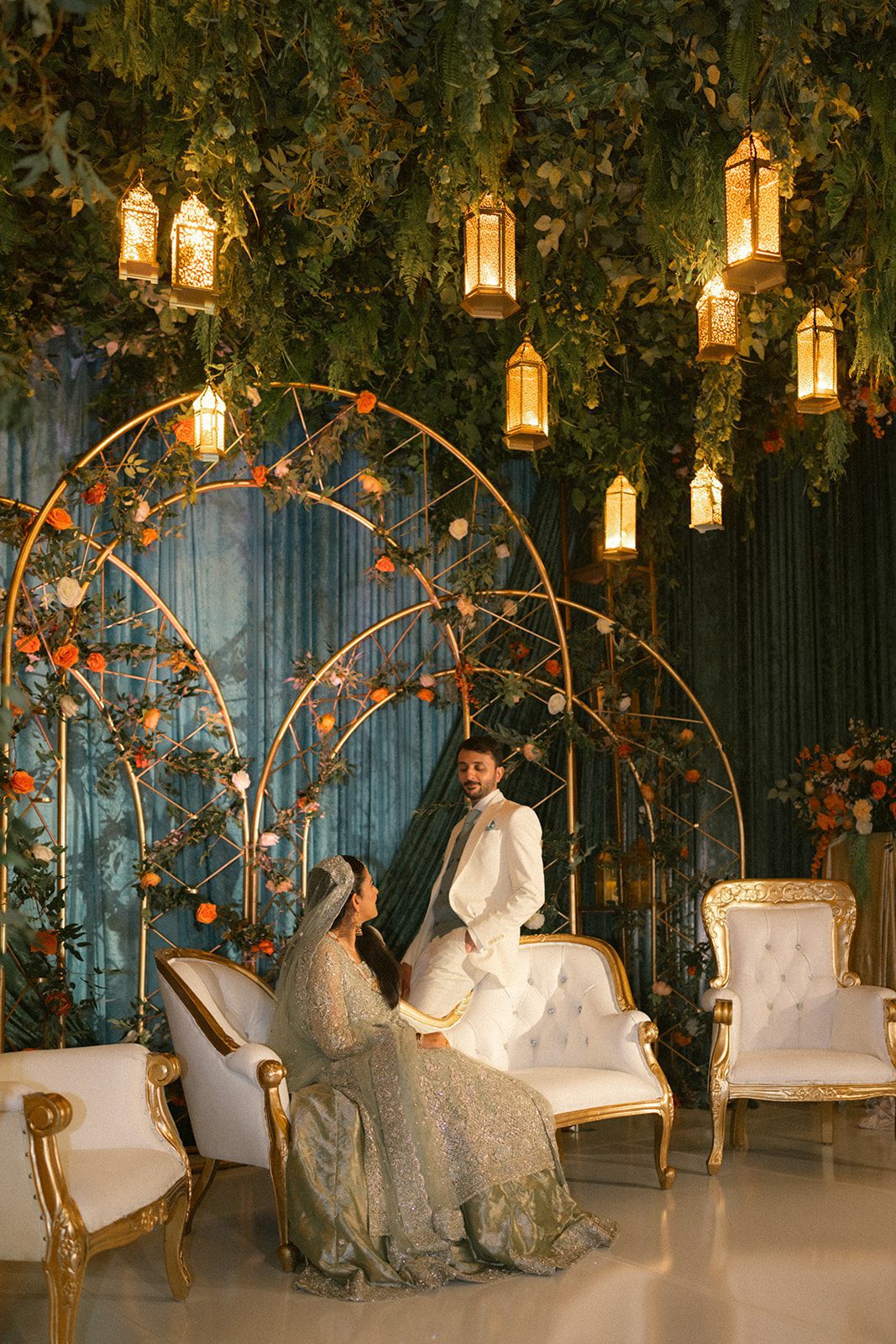 Couple posing on an ornate reception stage beneath hanging lanterns, lush greenery, and floral arches during their south asian wedding celebration.