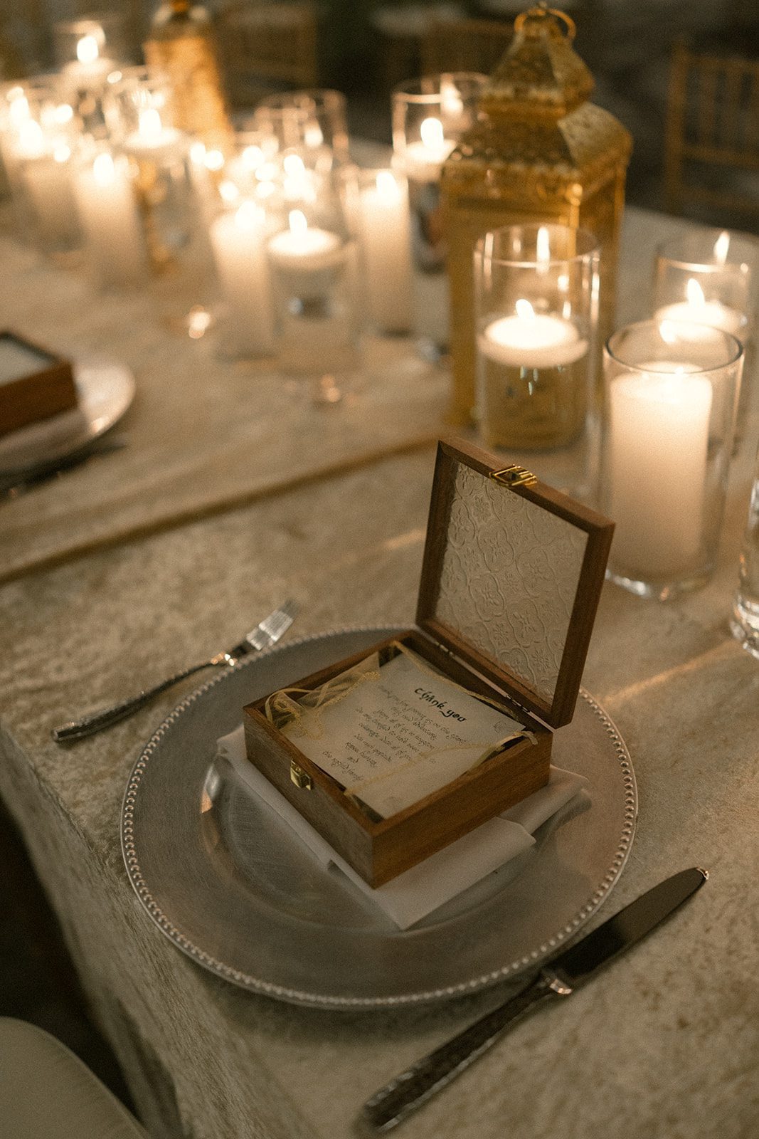 Candlelit indian wedding reception place setting featuring a wooden keepsake box, printed note, silver charger, and glowing gold lanterns.