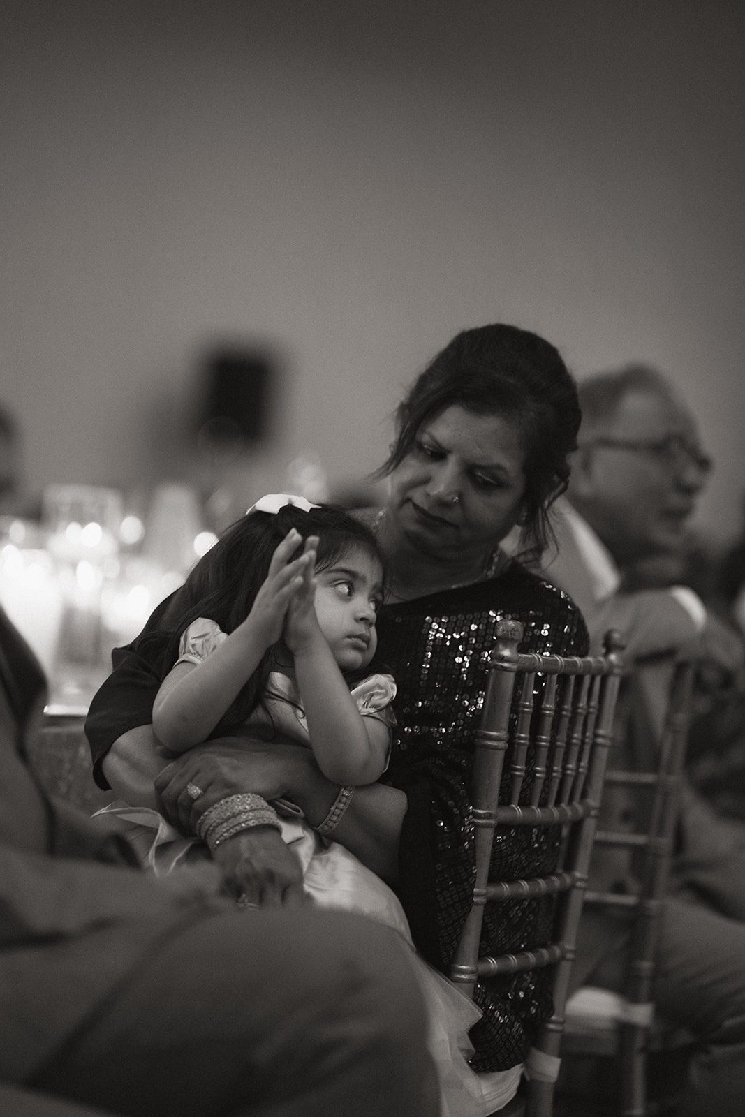 Black-and-white candid of a woman holding a young girl seated among guests at an indian wedding reception.