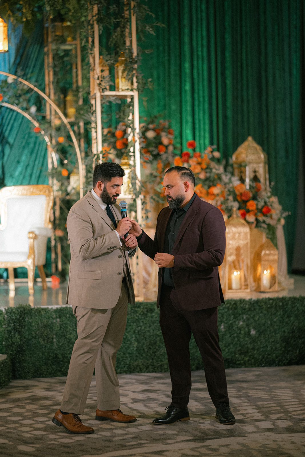 Two men speaking into a microphone in front of an elaborate green and floral stage at a south asian wedding reception.