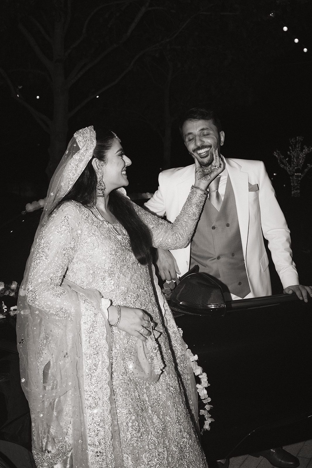 Black-and-white candid of the bride playfully touching the groom’s face beside a car after their multi day wedding celebration.
