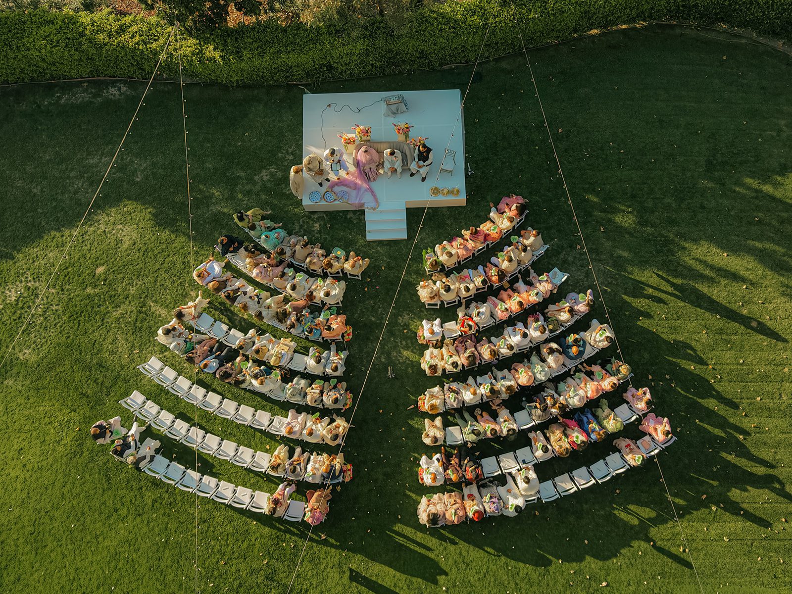 Aerial view of an outdoor multi day wedding ceremony with the couple and family seated on a white stage as guests gather in rows across the lawn.
