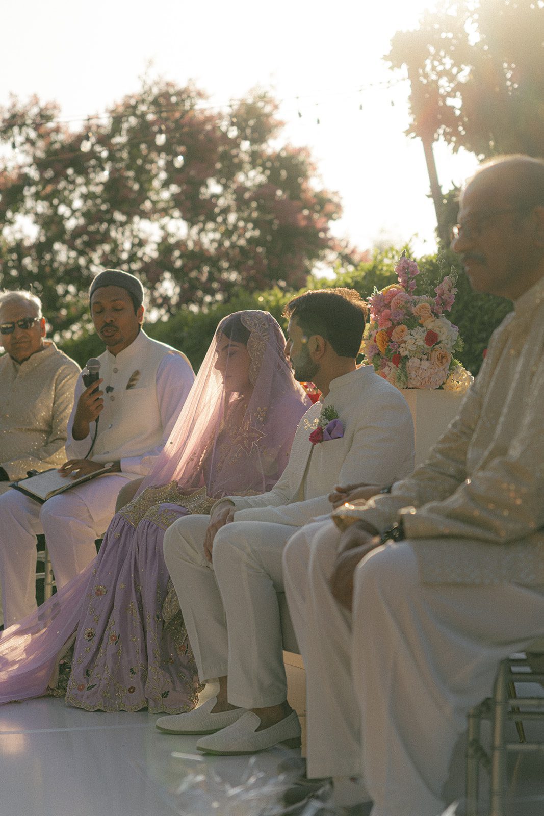 Bride and groom seated with family during an outdoor indian wedding ceremony, surrounded by floral arrangements in warm golden-hour light.