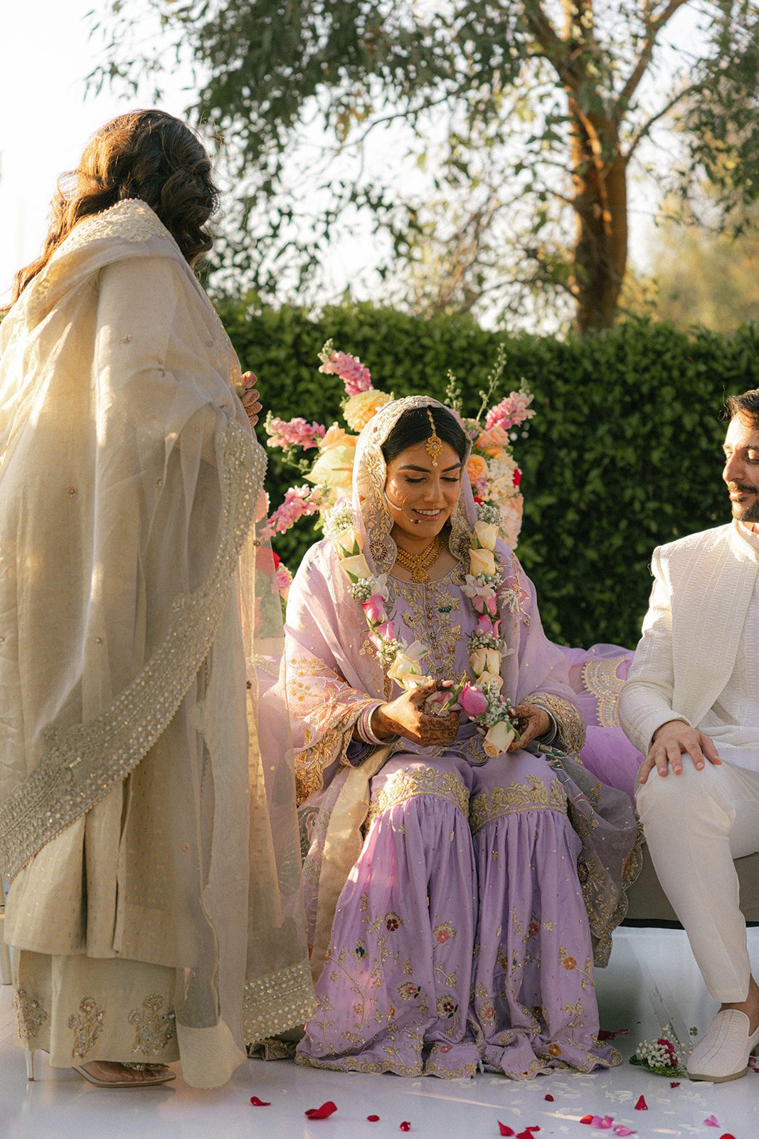 Bride in lavender traditional attire seated beside the groom during an outdoor indian wedding ceremony.