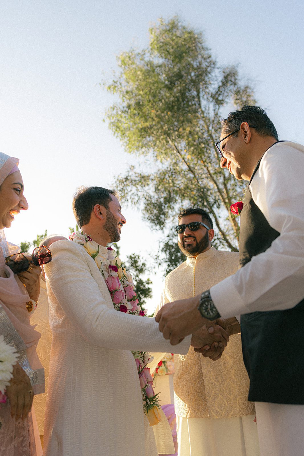 Groom greeting a guest with a handshake during a sunlit outdoor south asian wedding ceremony.