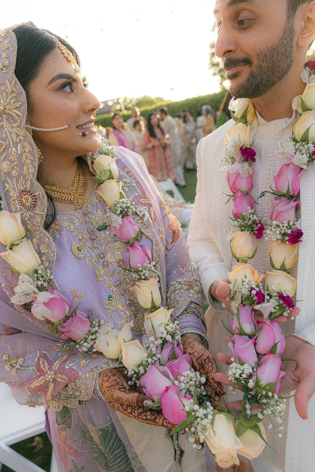 Close-up of the couple wearing pink and ivory rose garlands during their outdoor indian wedding ceremony, with the bride’s embroidered lavender attire and mehndi in view.