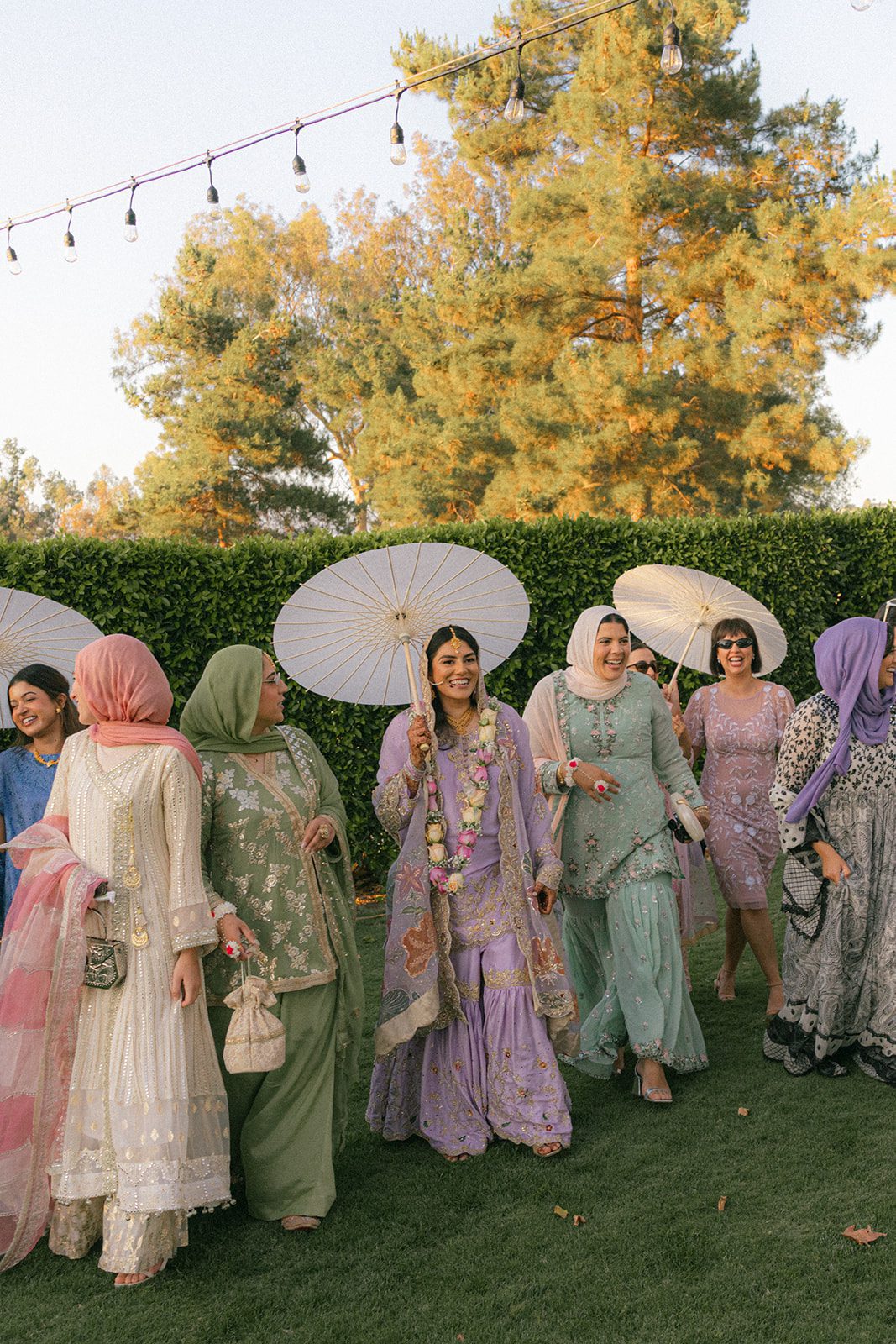 Bride in lavender wedding attire walking across the lawn with guests and white parasols during a colorful south asian wedding celebration.