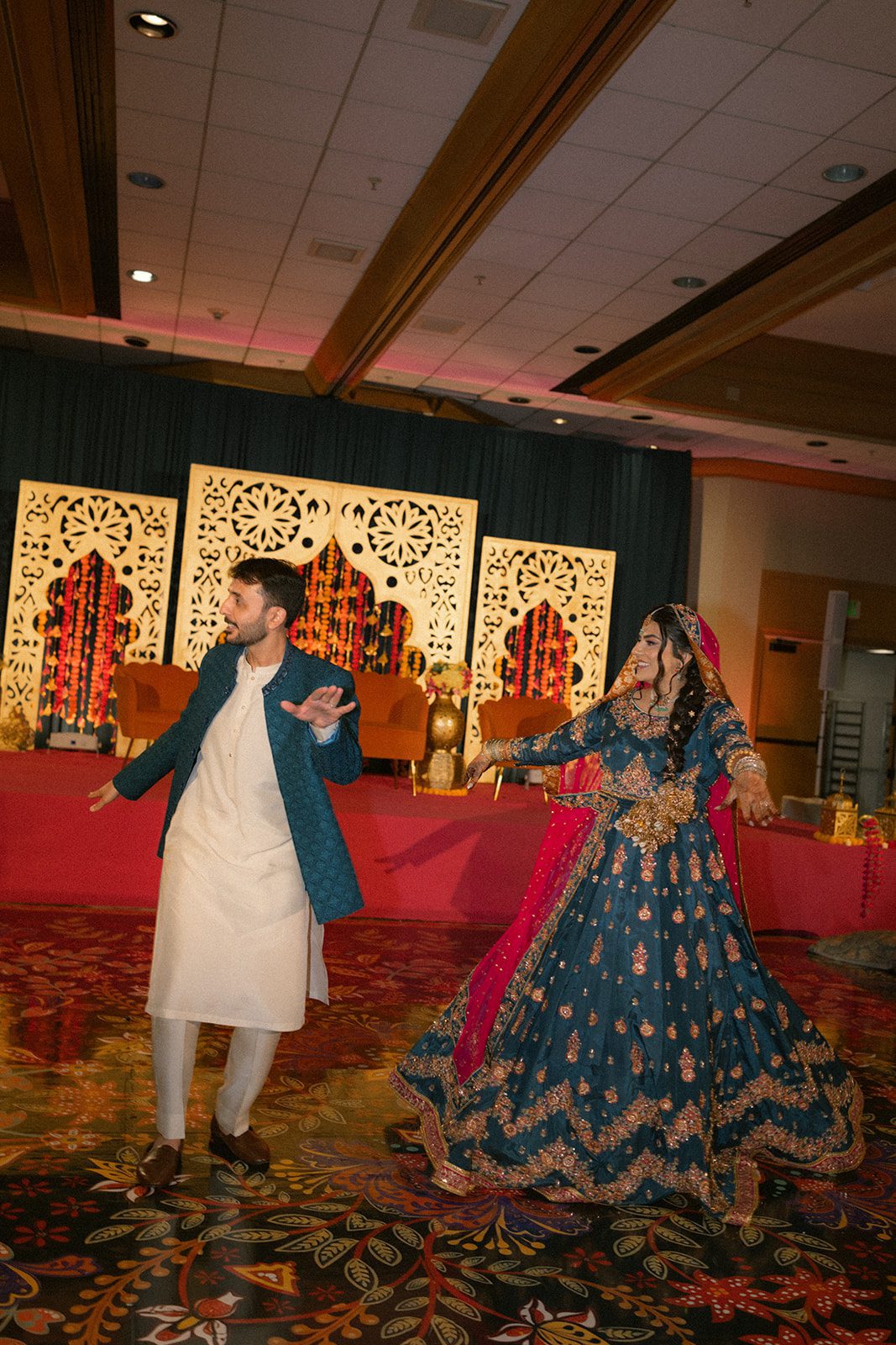 Couple dancing together in colorful traditional attire during a lively south asian wedding celebration with marigold decor and an ornate stage backdrop.