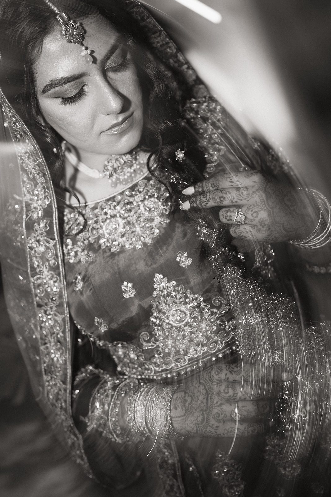 Black-and-white bridal portrait highlighting an embroidered veil, statement jewelry, bangles, and intricate mehndi during an indian wedding celebration.