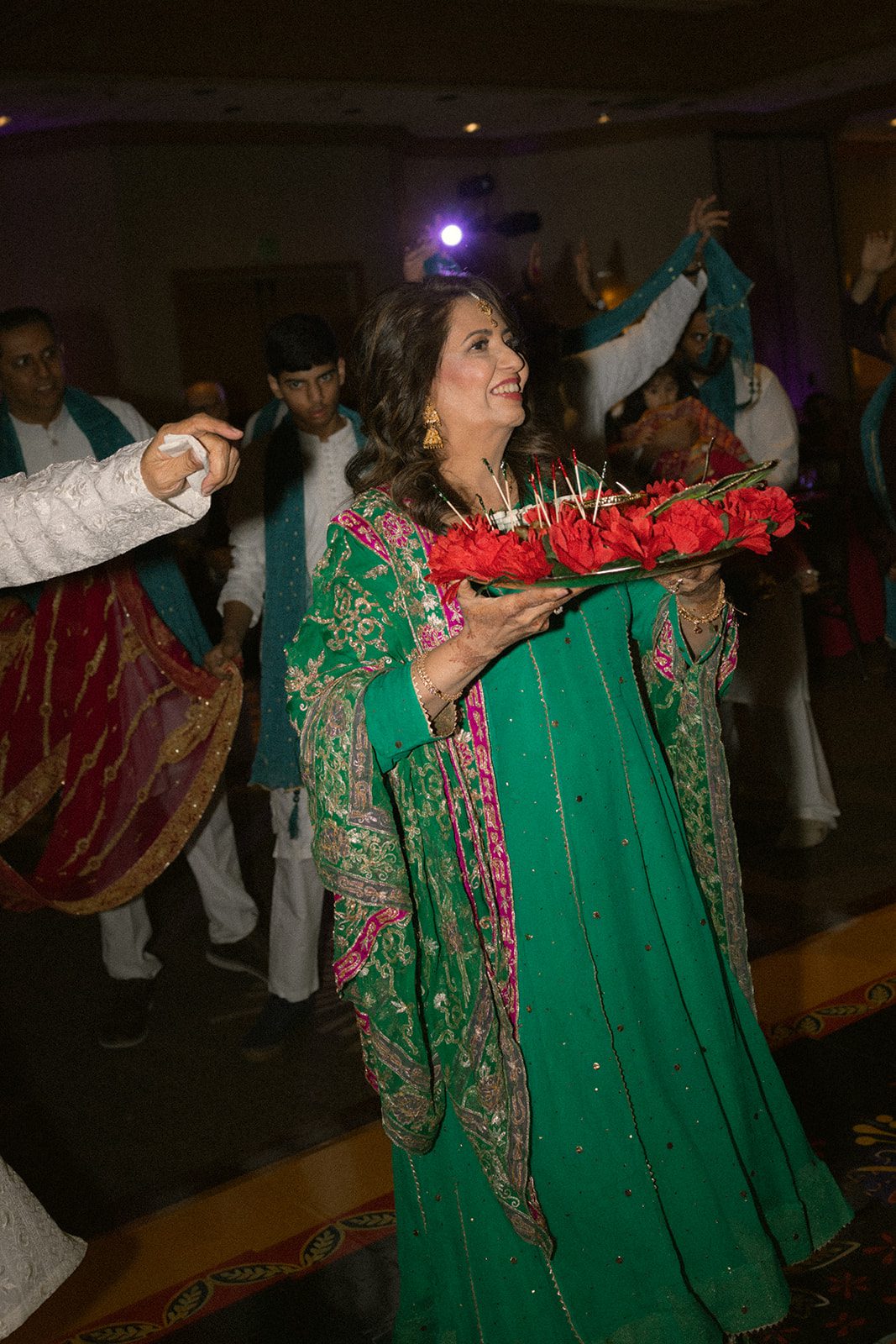 Woman in green traditional attire carrying a tray of red flowers during a lively indian wedding celebration.