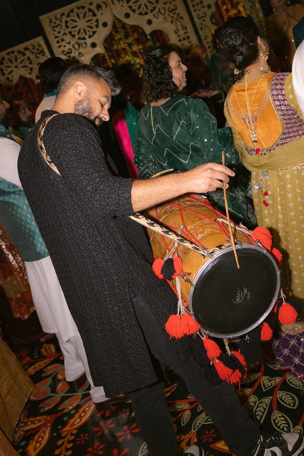 Dhol drummer performing among guests on the dance floor during a colorful south asian wedding event.