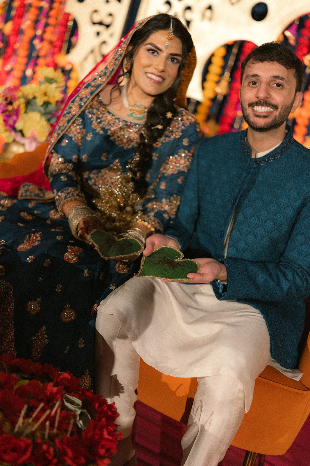Couple in coordinated blue traditional attire seated before colorful floral decor during a south asian wedding event, holding ceremonial leaves in their hands.