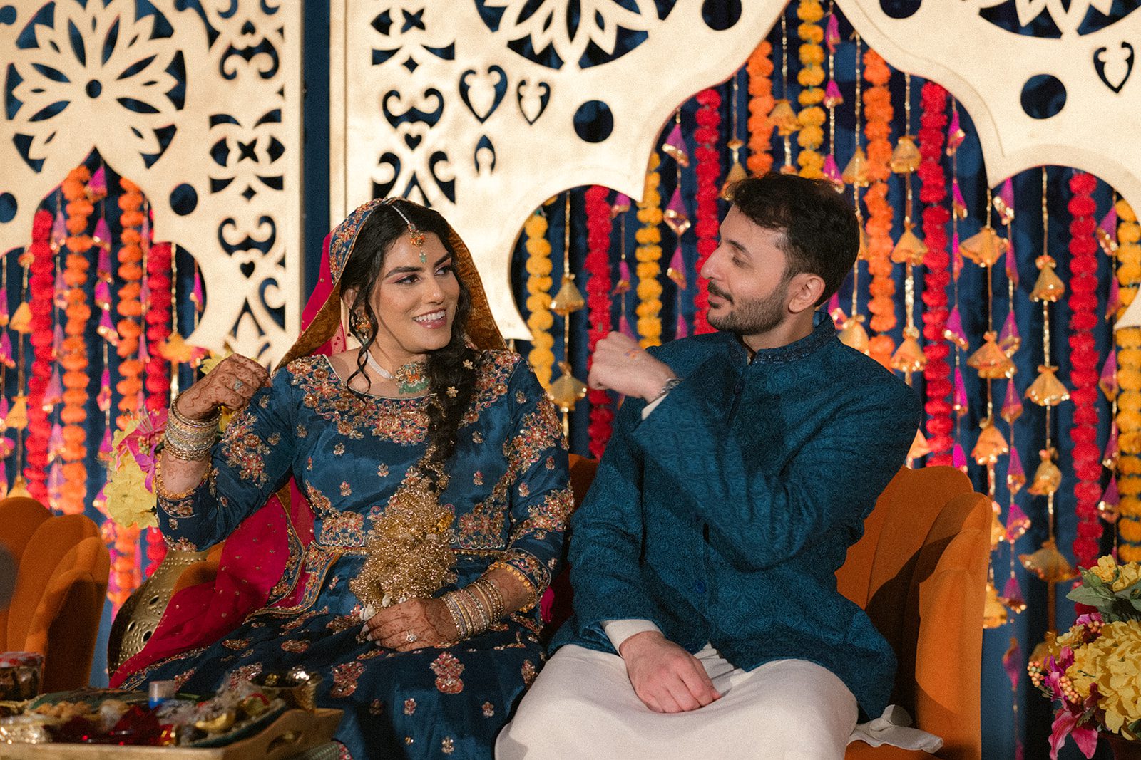 Bride and groom seated together in coordinated blue attire against vibrant marigold garlands during an indian wedding event, part of their multi day wedding celebration.