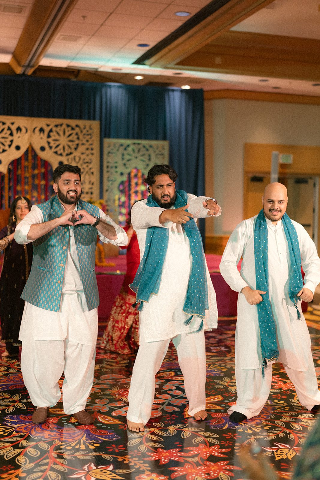 Three men in white traditional attire with teal accents performing a choreographed dance at a multi day wedding celebration.