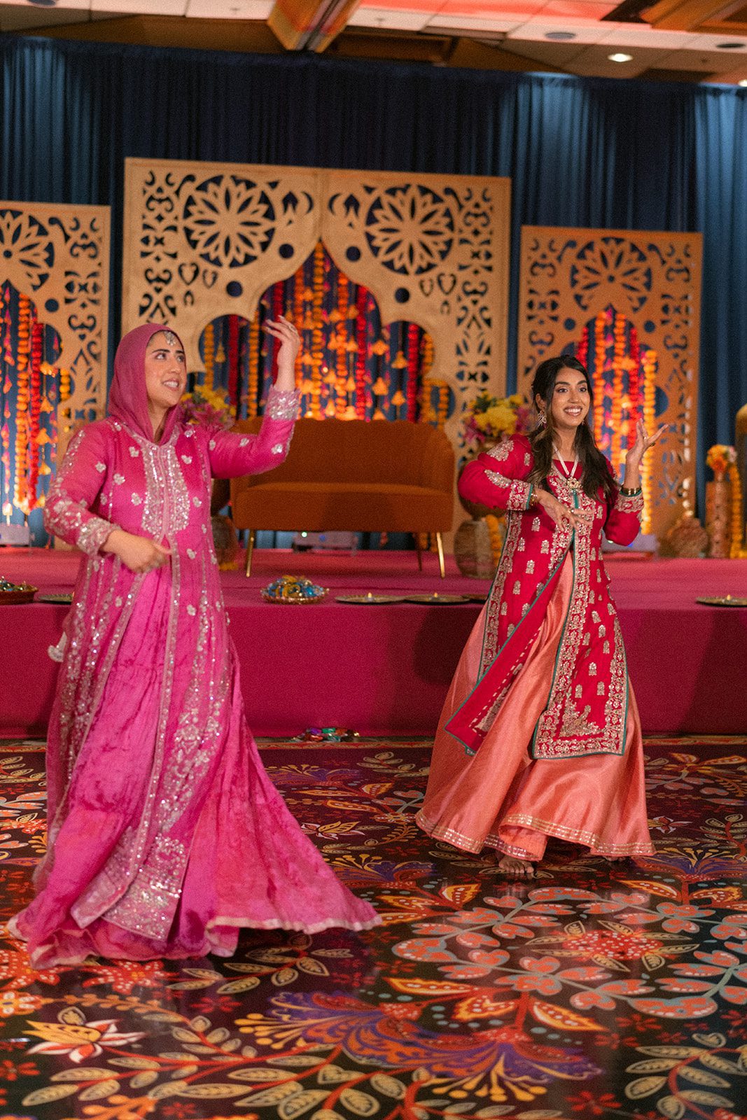  Two women in bright pink traditional outfits dancing in front of colorful floral decor during an indian wedding event.